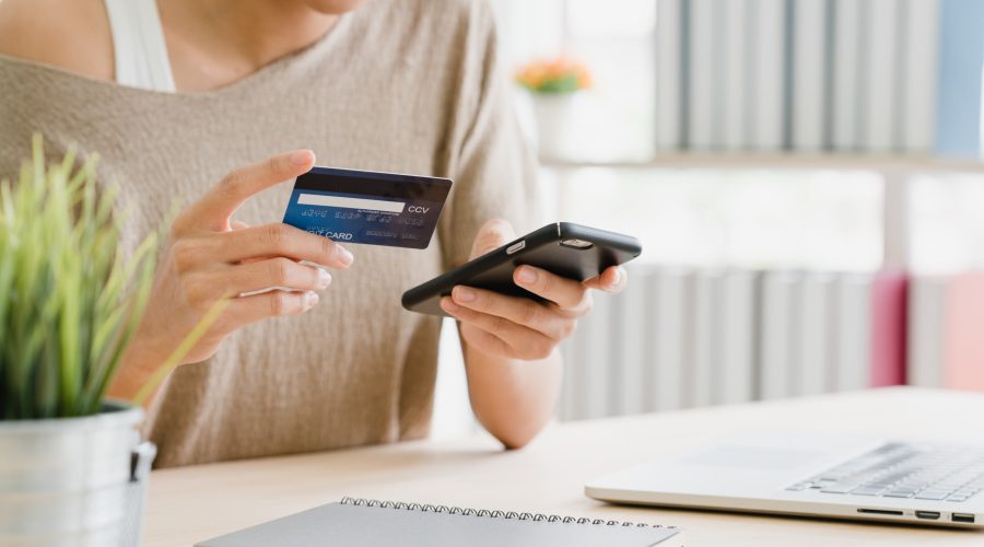 Beautiful Asian woman using smartphone buying online shopping by credit card while wear sweater sitting on desk in living room at home. Lifestyle woman at home concept.